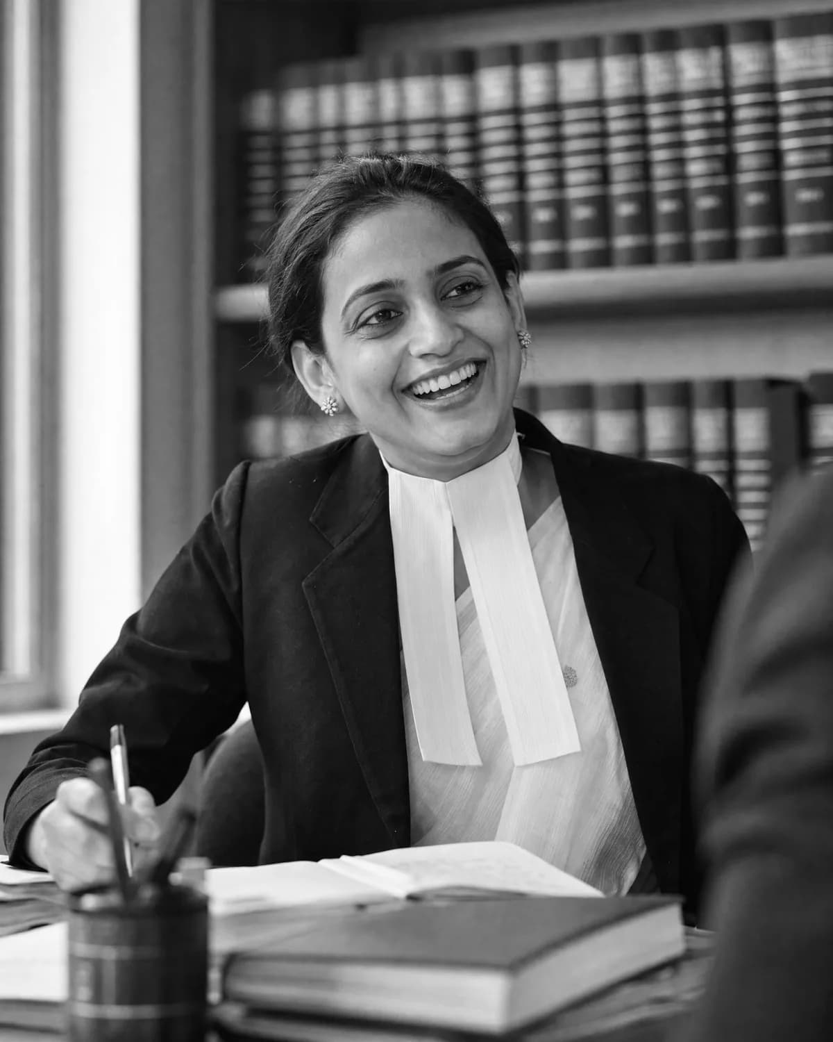 An advocate in a black coat and white bands laughing at her desk in chambers, law reports on the shelves behind her